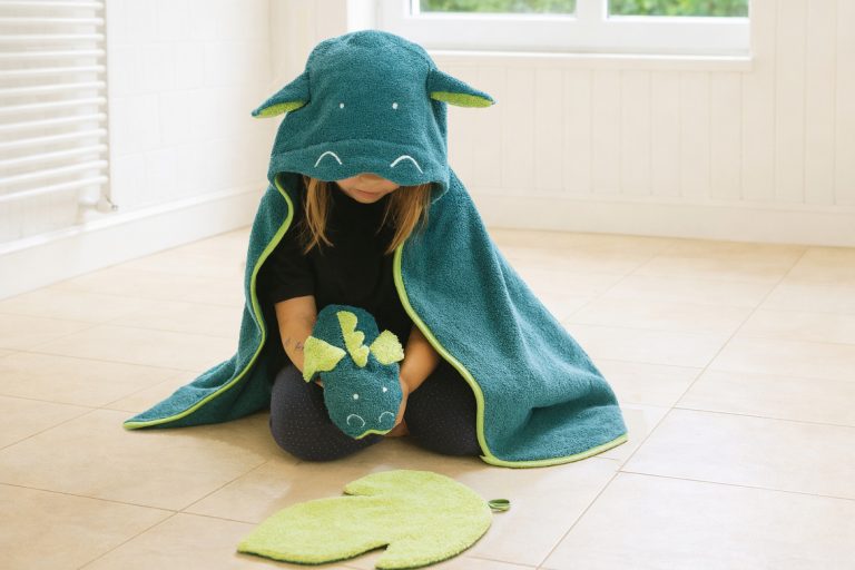 Kind eingekuschelt in ein grünes Drachenhandtuch, spielt mit einem Drachenwaschlappen und einem kleinen Handtuch in Form einer Flamme.